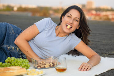 Glückliche junge Frau isst Käse, während sie beim Picknick auf dem Dach liegt - NGF00705