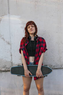Junge rothaarige Frau mit Kopfhörern hält Skateboard gegen die Wand - MGRF00047