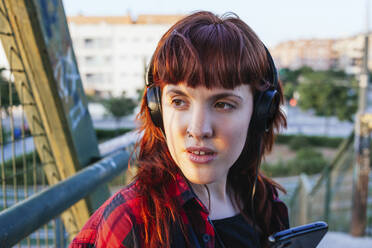 Nachdenkliche junge Frau, die wegschaut, während sie über Kopfhörer Musik hört - MGRF00052