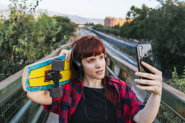 Rothaarige Frau mit Skateboard, die ein Selfie macht, während sie auf einer Fußgängerbrücke steht - MGRF00053