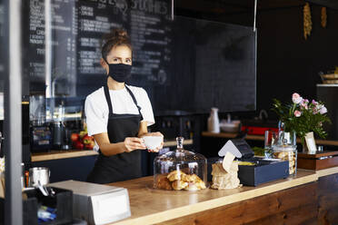 Porträt einer Kellnerin mit Gesichtsschutzmaske und Kaffeetasse bei der Arbeit in einem Café - BSZF01763