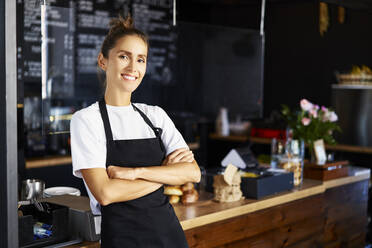 Porträt einer lächelnden Barista mit verschränkten Armen bei der Arbeit in einem Café - BSZF01766