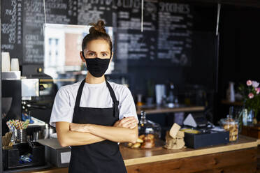 Porträt einer Kellnerin mit Gesichtsschutzmaske und verschränkten Armen in einem Cafe - BSZF01768