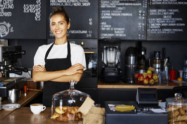 Porträt einer lächelnden Kellnerin mit verschränkten Armen, die in einem Café arbeitet - BSZF01779