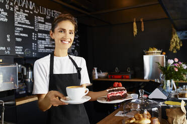 Porträt einer lächelnden Kellnerin, die Kaffee und Gebäck in einem Café serviert - BSZF01795