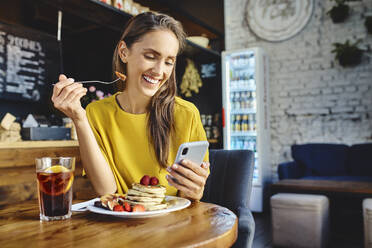 Lächelnde junge Frau, die eine Beere isst und dabei ihr Handy am Tisch im Café benutzt - BSZF01801
