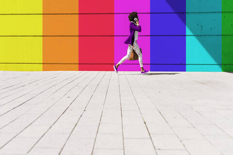 Young woman talking on smart phone while walking by colorful wall during sunny day stock photo