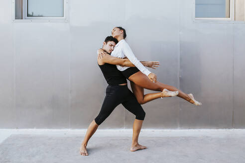 Young professional acrobats embracing each other while dancing against wall - MIMFF00270