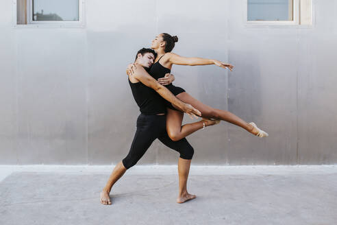 Young male and female professional acrobats embracing each other while dancing against wall - MIMFF00271