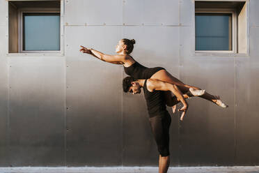 Male gymnast carrying female on shoulders while practicing dance posture by wall - MIMFF00273