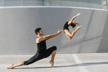 Female gymnast doing acrobat while male dancer practicing stag leap by gray wall - MIMFF00279