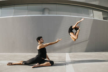 Female dancer doing acrobat while male gymnast practicing stag leap by gray wall - MIMFF00280