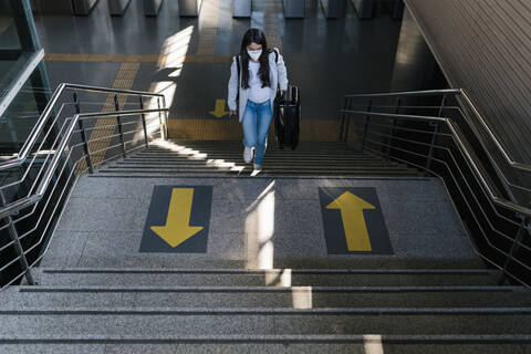 Woman carrying luggage while moving up on staircase at railroad station during pandemic stock photo