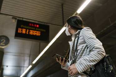 Woman looking away while standing with smart phone waiting at railroad station - EGAF01003