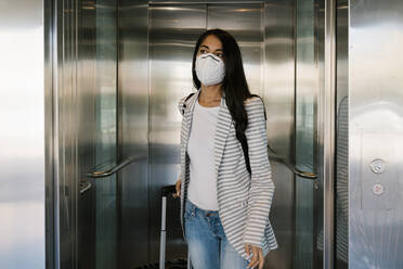 Woman wearing protective face mask walking out with luggage from elevator at station - EGAF01006