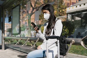 Frau wartet auf den Bus und benutzt ihr Smartphone an einem sonnigen Tag während der COVID-19-Pandemie - EGAF01008