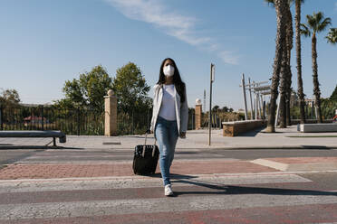 Frau mit Gepäck überquert Straße gegen blauen Himmel an einem sonnigen Tag während der Pandemie - EGAF01012