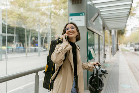 Smiling woman talking on mobile phone while standing at tram station in city stock photo