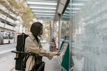 Frau mit Instrumentenkoffer, die einen Fahrkartenautomaten benutzt, während sie mit einem Elektroroller an einer Straßenbahnhaltestelle in der Stadt steht - VABF03968