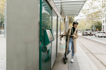 Frau mit Fahrradhelm, die mit einem Elektroroller an einer Straßenbahnhaltestelle in der Stadt wartet - VABF03974