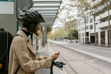 Mittlere erwachsene Frau mit Instrumentenkoffer und Elektroroller, die ein Mobiltelefon benutzt, während sie an einer Straßenbahnhaltestelle in der Stadt steht - VABF03976