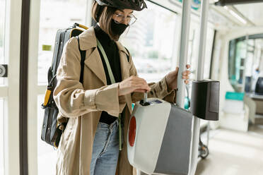 Frau mit Gesichtsmaske und Fahrradhelm, die in der Straßenbahn eine Fahrkarte in den Entwertungsautomaten steckt - VABF03977