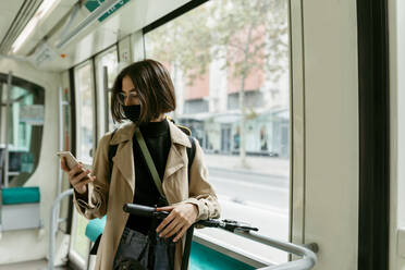 Frau mit Elektroroller und Gesichtsmaske, die in der Straßenbahn steht und ein Mobiltelefon benutzt - VABF03980