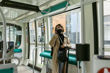 Frau mit Elektroroller und Gesichtsmaske, die in der Straßenbahn mit ihrem Handy telefoniert - VABF03981