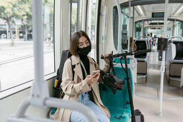 Mittlere erwachsene Frau mit Gesichtsmaske, die ein Mobiltelefon benutzt, während sie mit einem Elektroroller in der Straßenbahn sitzt - VABF03982