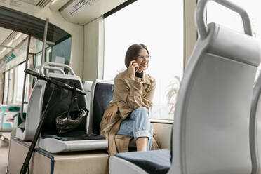 Lächelnde Frau, die mit einem Mobiltelefon spricht, während sie mit einem elektrischen Roller in der Straßenbahn sitzt - VABF03988