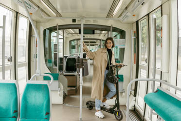 Lächelnde Frau mit Elektro-Scooter in der Straßenbahn stehend - VABF03991