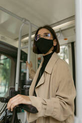 Mittlere erwachsene Frau mit Gesichtsmaske, die mit einem Tretroller in der Straßenbahn steht - VABF03993