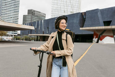 Lächelnde Frau mit Fahrradhelm, die wegschaut, während sie mit einem Elektroroller in der Stadt spazieren geht - VABF03997