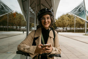 Smiling woman wearing cycling helmet standing with electric push scooter on footpath - VABF04003