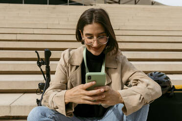 Frau lächelt, während sie ein Mobiltelefon benutzt, während sie mit einem elektrischen Tretroller auf einer Treppe sitzt - VABF04022