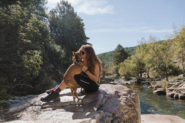 Frau küsst Hund, während sie auf einem Felsen im Wald bei La Pedriza, Madrid, Spanien, sitzt - MRRF00650