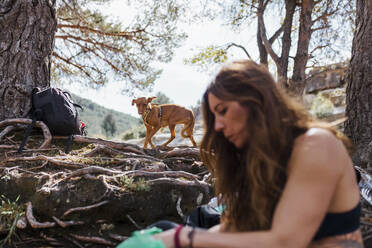 Mittlere erwachsene Frau sitzend mit Hund im Hintergrund im Wald La Pedriza, Madrid, Spanien - MRRF00675