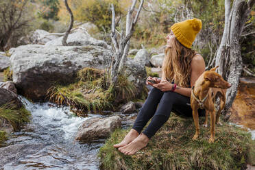 Frau, die ein Mobiltelefon benutzt, während sie mit ihrem Hund auf einem Felsen im Wald bei La Pedriza, Madrid, Spanien, sitzt - MRRF00688