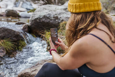 Trekker taking photo of river through mobile phone while sitting in forest at La Pedriza, Madrid, Spain - MRRF00689