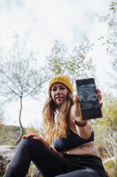Mid adult woman showing mobile phone while sitting in forest at La Pedriza, Madrid, Spain - MRRF00692