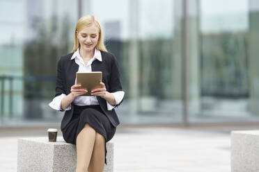 Blonde Geschäftsfrau benutzt ein digitales Tablet, während sie auf einer Bank vor einem Gebäude sitzt - GGGF00083