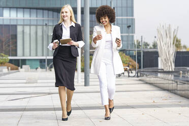 Lächelnde Geschäftsfrauen, die ein digitales Tablet und ein Smartphone benutzen, während sie gegen ein Gebäude laufen - GGGF00096