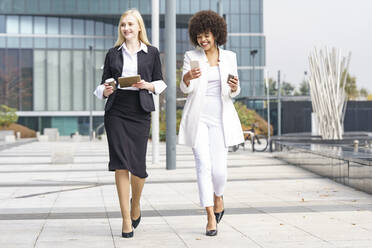 Smiling businesswomen using digital tablet and smart phone while walking against building - GGGF00096