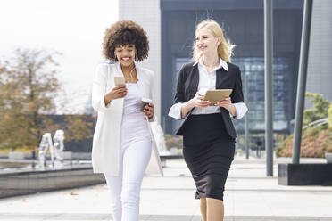 Smiling businesswomen walking with smart phone and digital tablet on footpath - GGGF00102