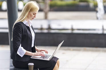 Blond businesswoman working on laptop while sitting on bench - GGGF00103