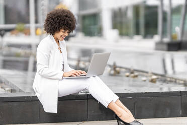 Lächelnde Geschäftsfrau, die an einem Laptop arbeitet, während sie auf einer Bank im Freien sitzt - GGGF00107