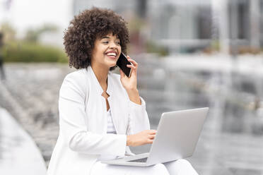 Lächelnde Geschäftsfrau, die auf einer Bank im Freien sitzt und mit ihrem Smartphone spricht - GGGF00111