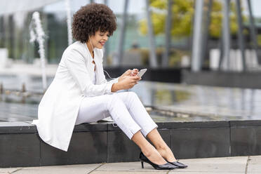 Geschäftsfrau mit In-Ear-Kopfhörern, die ein Mobiltelefon benutzt, während sie auf einer Bank im Freien sitzt - GGGF00116
