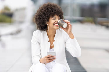 Smiling businesswoman with mobile phone drinking coffee while sitting at city - GGGF00120