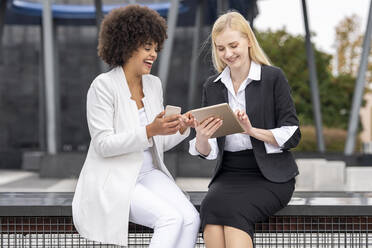 Smiling businesswomen using mobile phone and digital tablet while sitting outdoors - GGGF00121